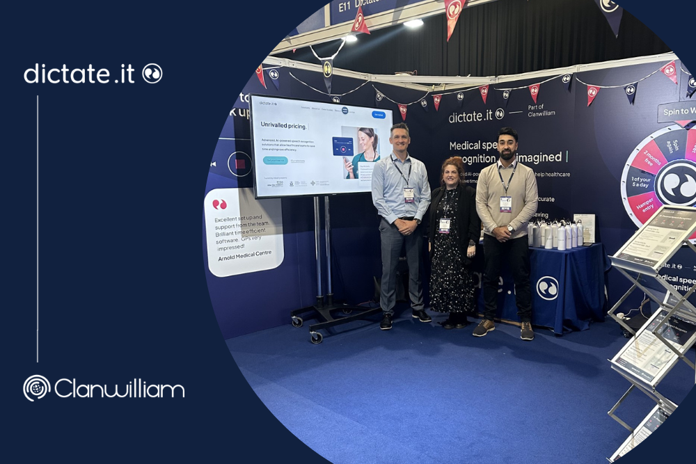 The Dictate.IT Primary Care team at the RCGP Annual Conference 2023. The image features a large TV screen with the Dictate.IT website on it and 3 people (2 men and 1 woman) stood in front of a table with some white water bottles on it.